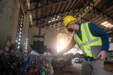 Worker in a safety vest and mask inspects a pile of plastic bottles within a large recycling facility